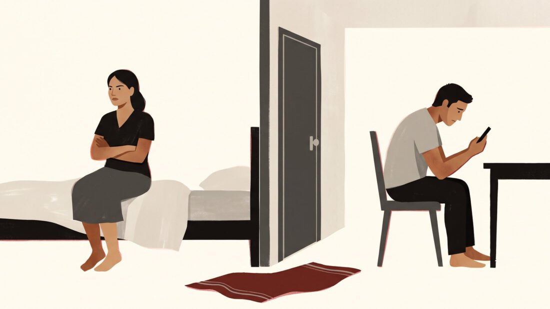 A couple sit back to back on either side of a wall, she on the bed with arms crossed, he on his phone, a red towel on the floor.